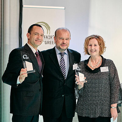 Three colleagues stand in front of the "Fair'n Green" banner and hold up two "Fair'n Green" trophies