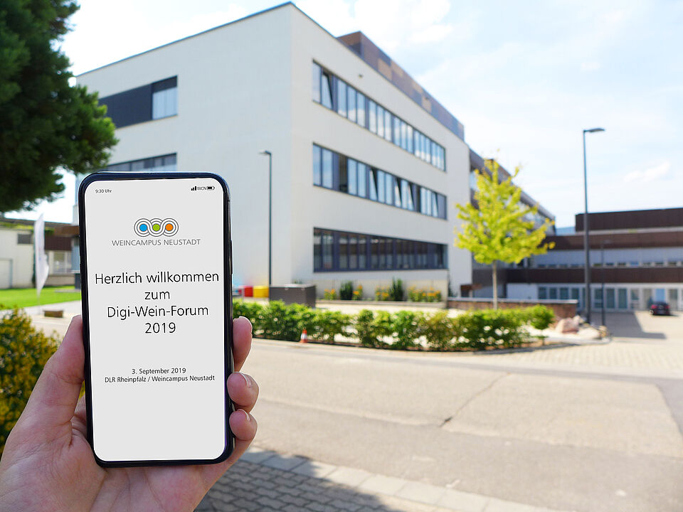 Handy Screen mit der Aufschrift: "Herzlich Willkommen zum Digi- Weinforum 2019" vor dem Gebäude des Weincampus Neustadt